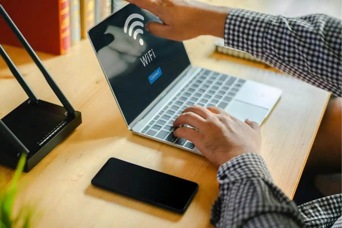 Gros plan sur des mains d'homme assis à un bureau, en train de connecter son ordinateur portable au réseau wifi de sa résidence étudiante. Sur le bureau sont également posés un smartphone, un modem et quelques livres.