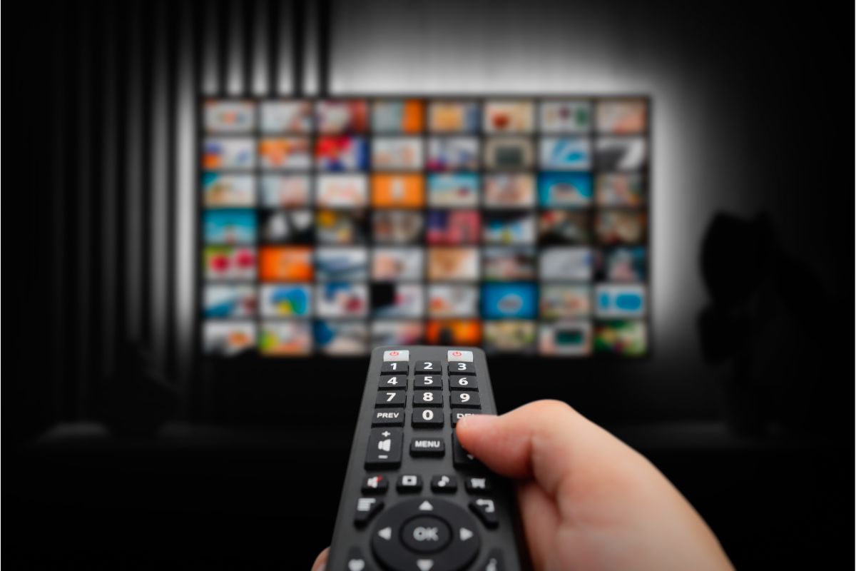 Main tenant une télécommande pointée vers un téléviseur affichant un mur de vignettes colorées représentant plusieurs chaînes ou contenus multimédias en streaming