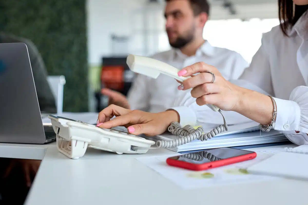 Collaboratrice décrochant un poste téléphonique IP, smartphone posé sur le bureau Collaboratrice décrochant un poste téléphonique IP, smartphone posé sur le bureau